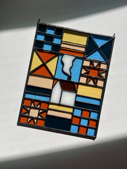Stained glass textile inspired panel depicting a home in the center in shades of teal, pink, white, blue, and yellow, against a white background with bright sunlight across