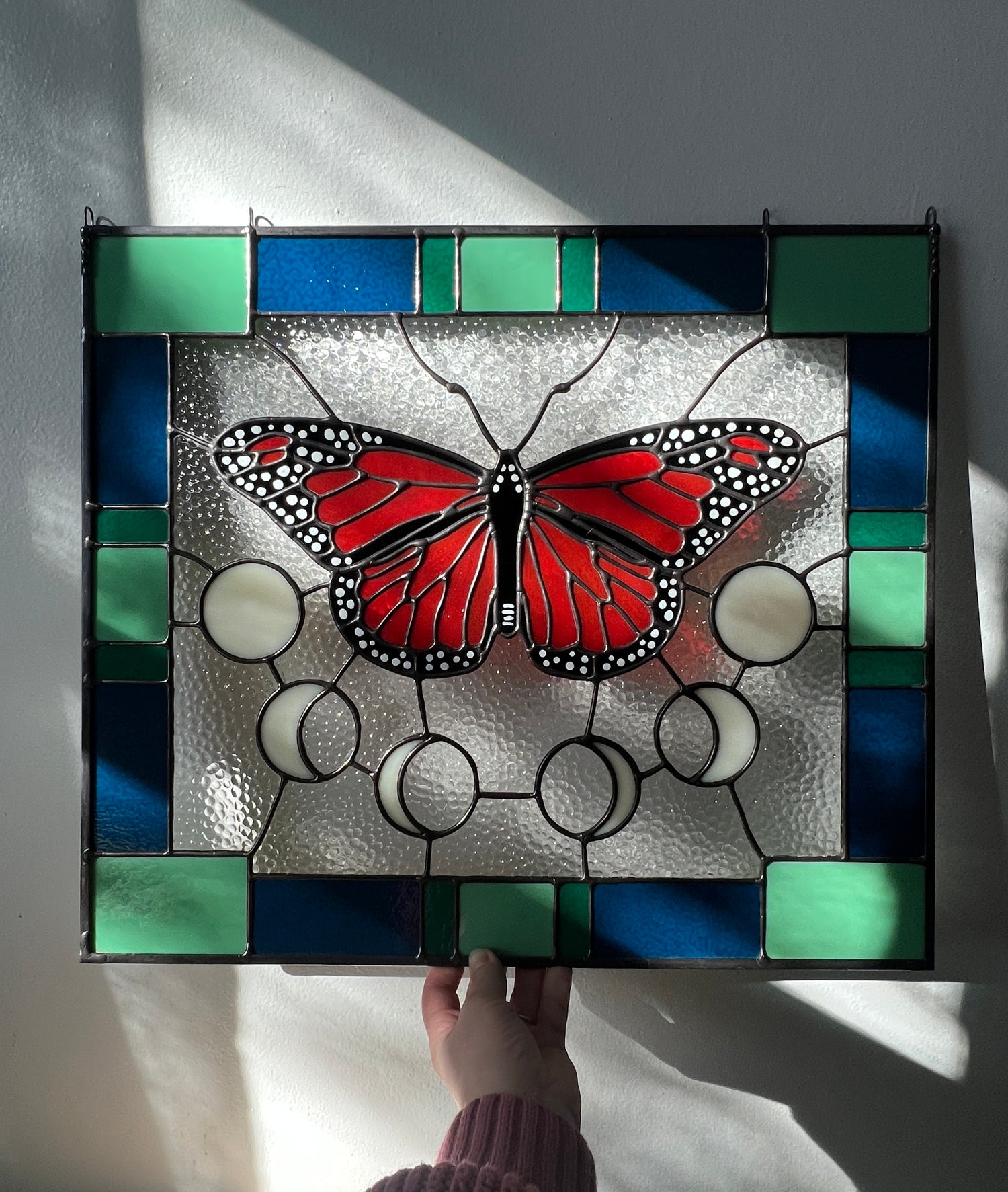 A large stained glass panel with a thick blue and green geometric border and a monarch butterfly and moon phases in the center, with textured clear glass in the background. The panel is held by a hand against a white wall with soft sunlight flowing across the piece.