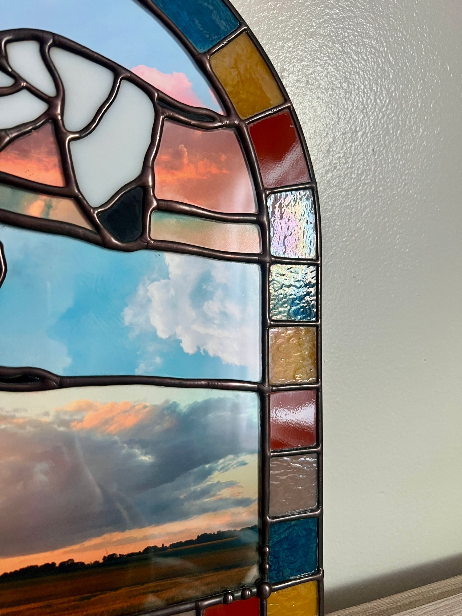Close up of stained glass panel with a colorful border featuring two birds and a sunset scene on a white wall.