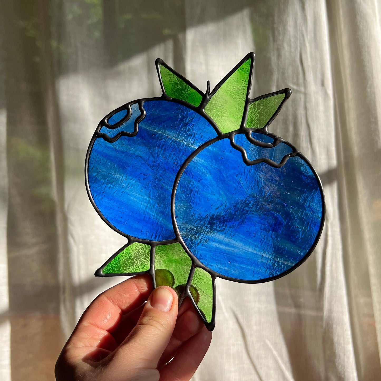 hand holding a textured blue stained glass suncatcher depicting a small bunch of blueberries with leaves in front of a window