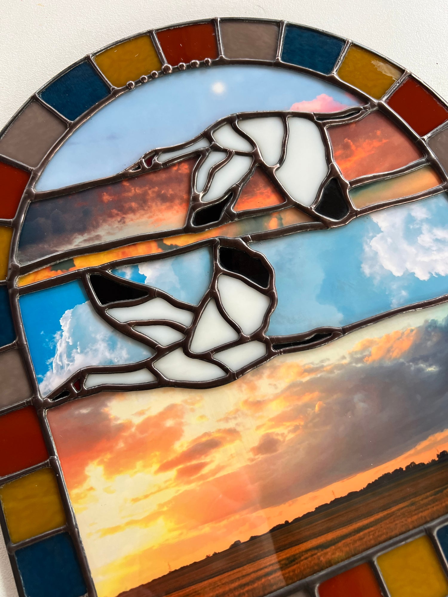 Close up image of a stained glass panel with a landscape design created by collaged photographs and two cranes flying