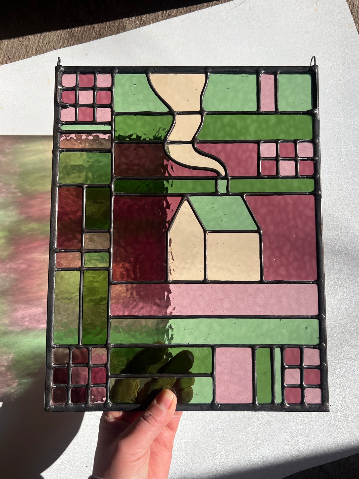Stained glass panel with geometric design held by a hand on a white surface, with bright sunlight casting colorful refractions