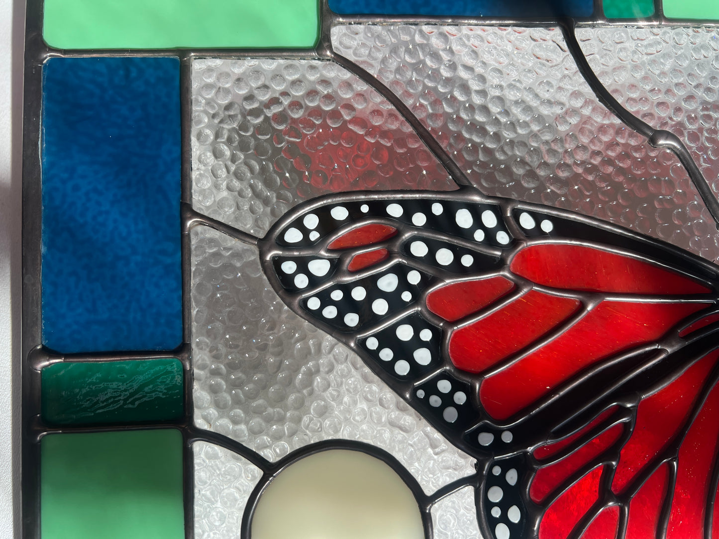Close up of a large stained glass panel with a thick blue and green geometric border and a monarch butterfly and moon phases in the center, with textured clear glass in the background. Photo highlights the hand painted white spots of the monarch's wing and the smooth solder.
