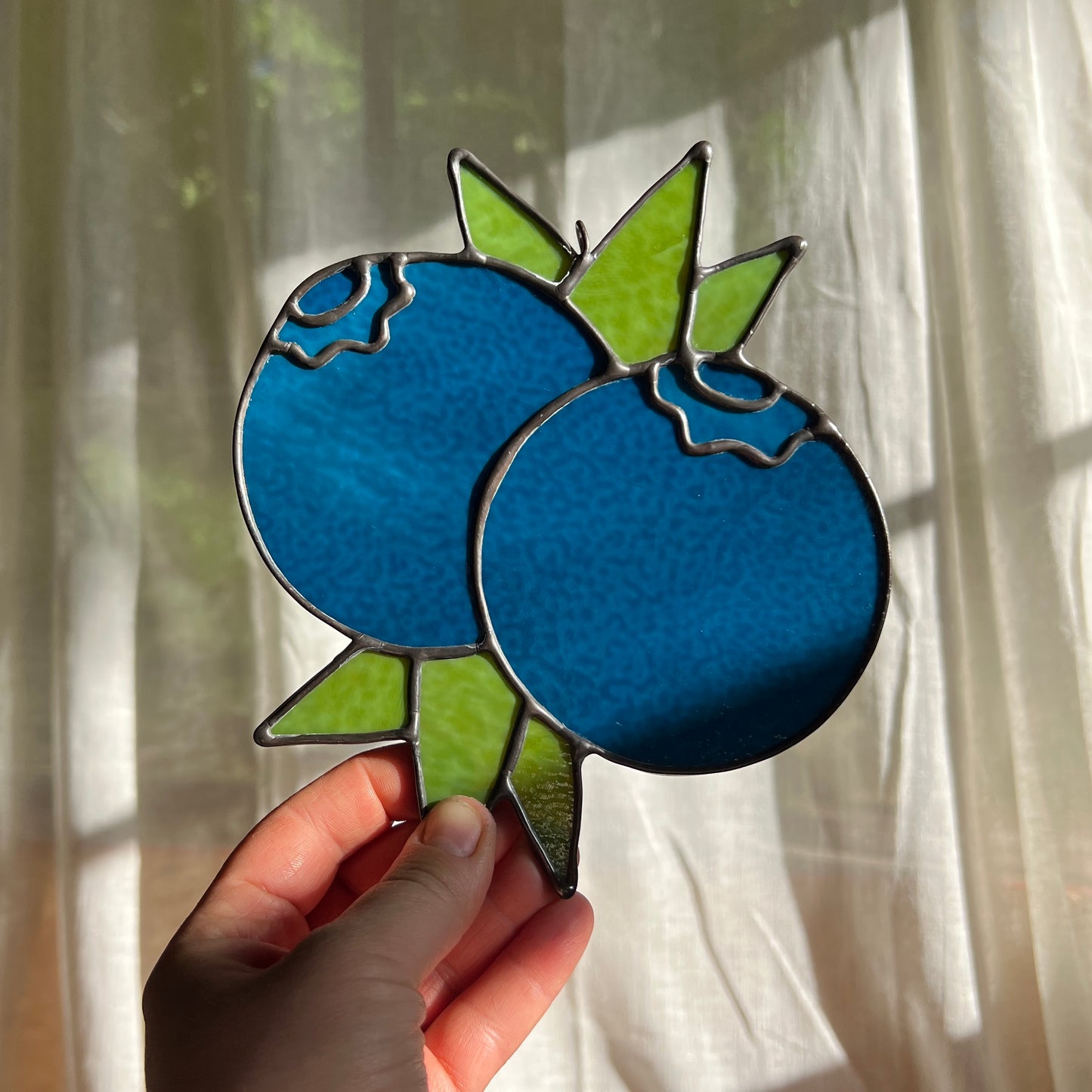 hand holding a dark blue stained glass suncatcher depicting a small bunch of blueberries with leaves in front of a window