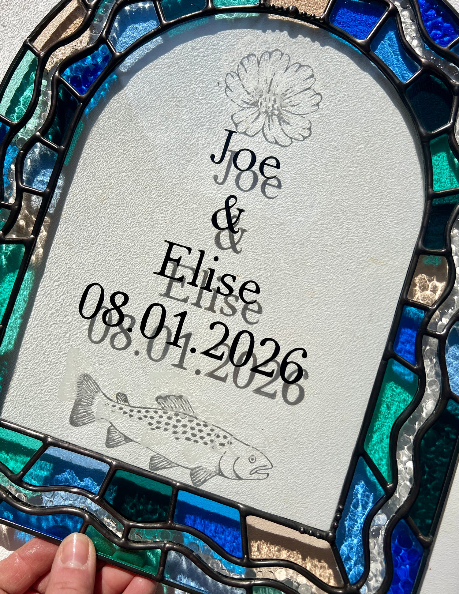 Slight close up of a stained glass panel in the shape of an arch with a multicolor blue, teal, turquoise, and pink border. Within the border is squiggle detail in clear textured glass. The center of the arch is clear uncolored glass, with personalized text and hand etched details of a cosmos flower and trout. The piece is held by a hand against a white wall, with sunlight casting refractions and shadows of the text, colored glass, and etchings.