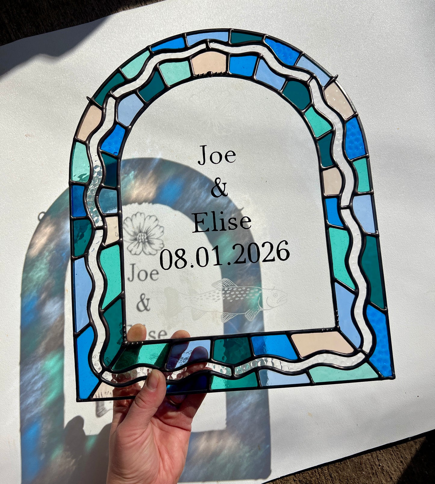 Stained glass panel in the shape of an arch with a multicolor blue, teal, turquoise, and pink border. Within the border is squiggle detail in clear textured glass. The center of the arch is clear uncolored glass, with personalized text and hand etched details of a cosmos flower and trout. The piece is held by a hand against a white wall, with sunlight casting refractions and shadows of the text, colored glass, and etchings.