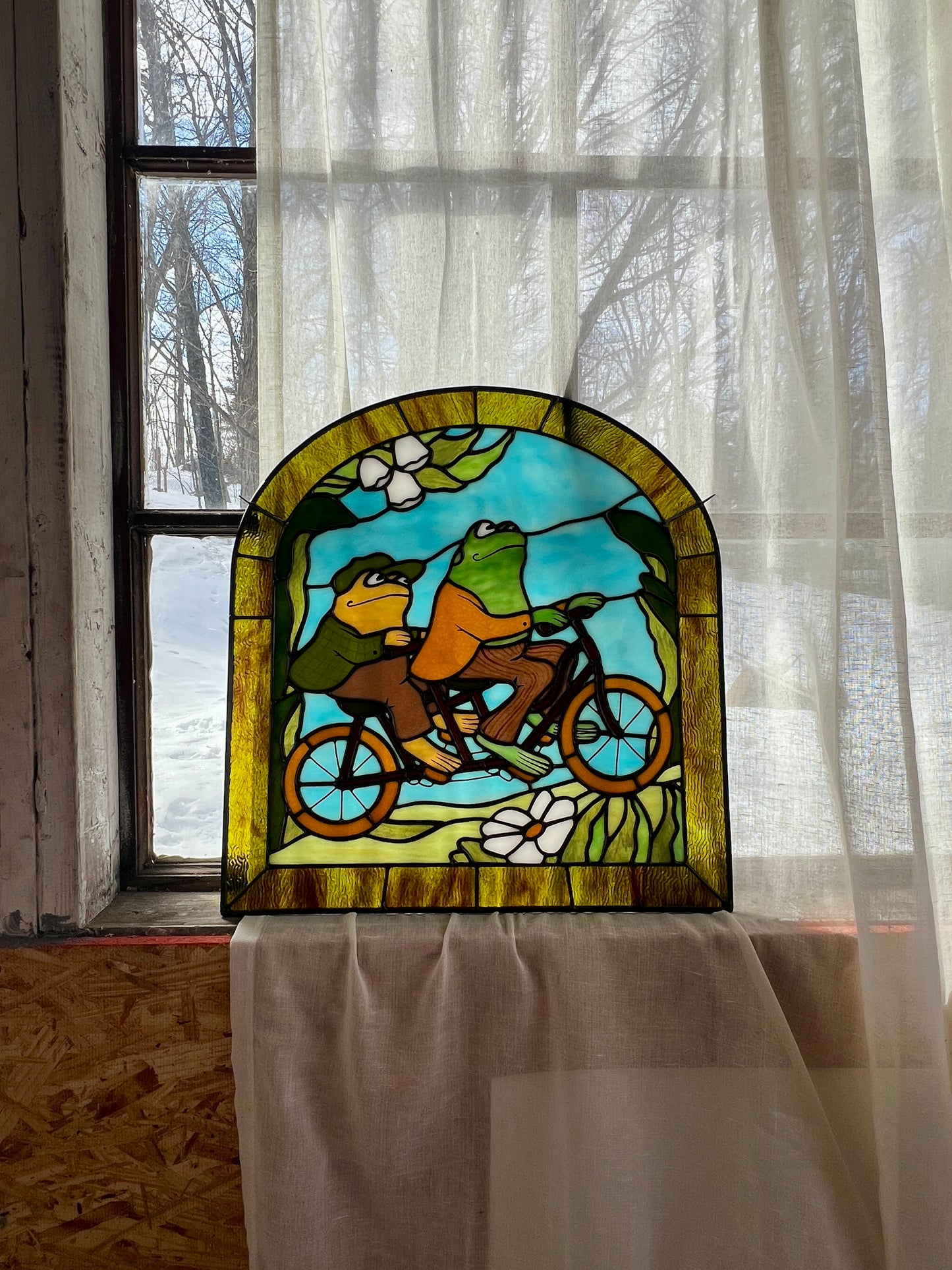 stained glass panel depicting frog and toad riding a bicycle resting in a window against a sheer curtain with a snowy scene outside