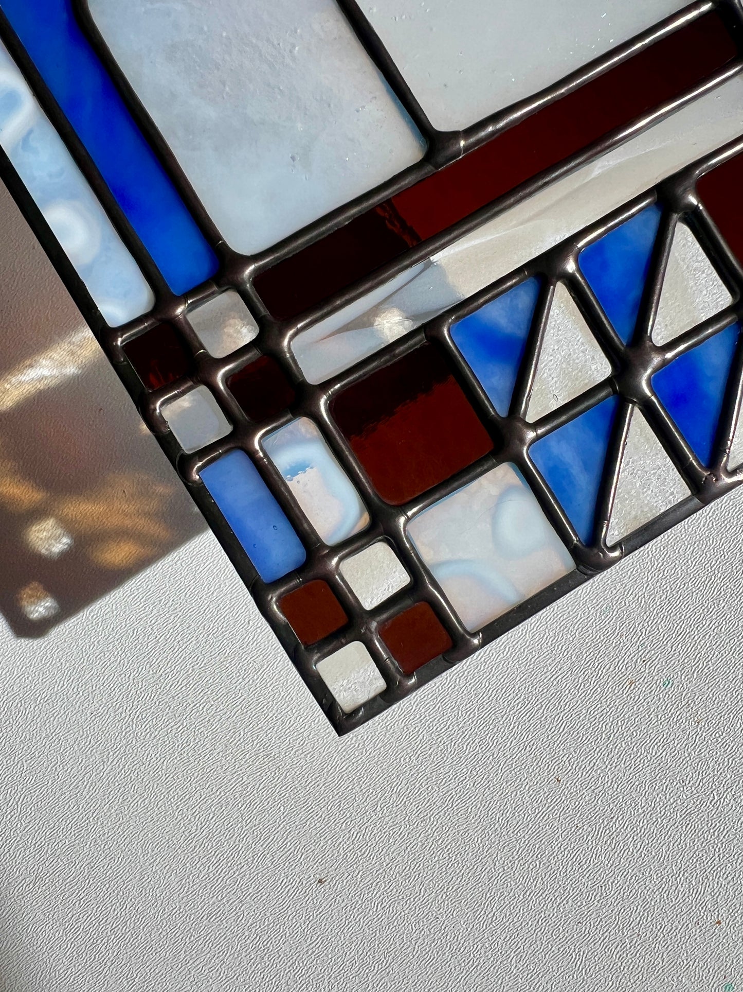 Close up of a textile inspired stained glass piece highlighting glass in various shades of blue, white, and brown, and smooth solder details held above a white surface