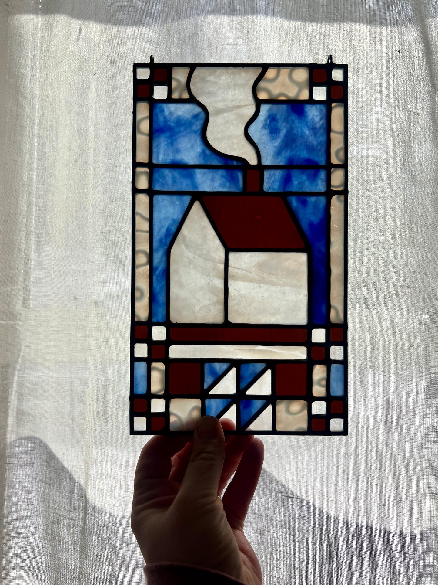 Stained glass textile inspired panel depicting a home in the center in shades of blue, brown, and white, held by a hand in front of a brightly backlit sheer white curtain
