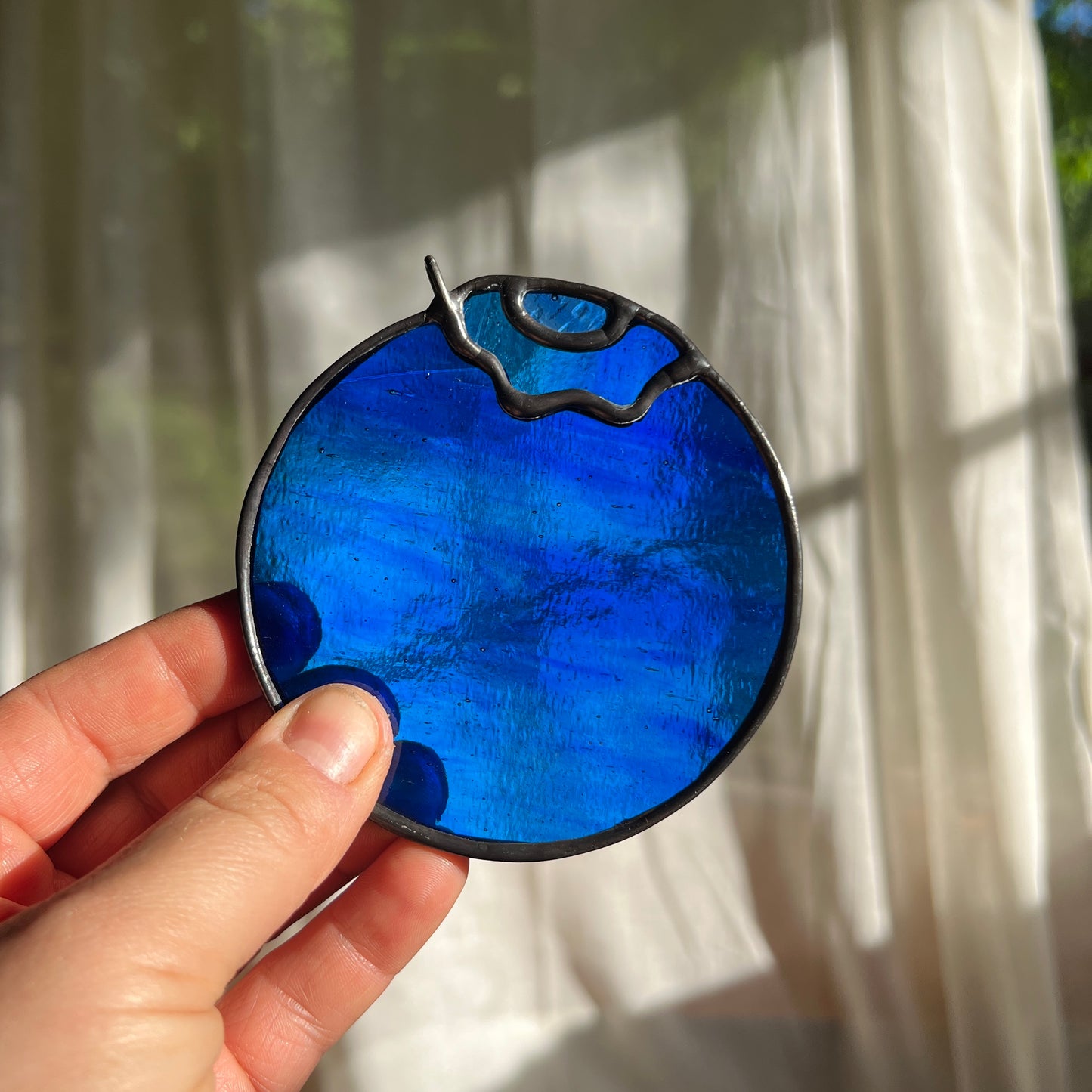 hand holding a wispy blue stained glass blueberry in front of a window