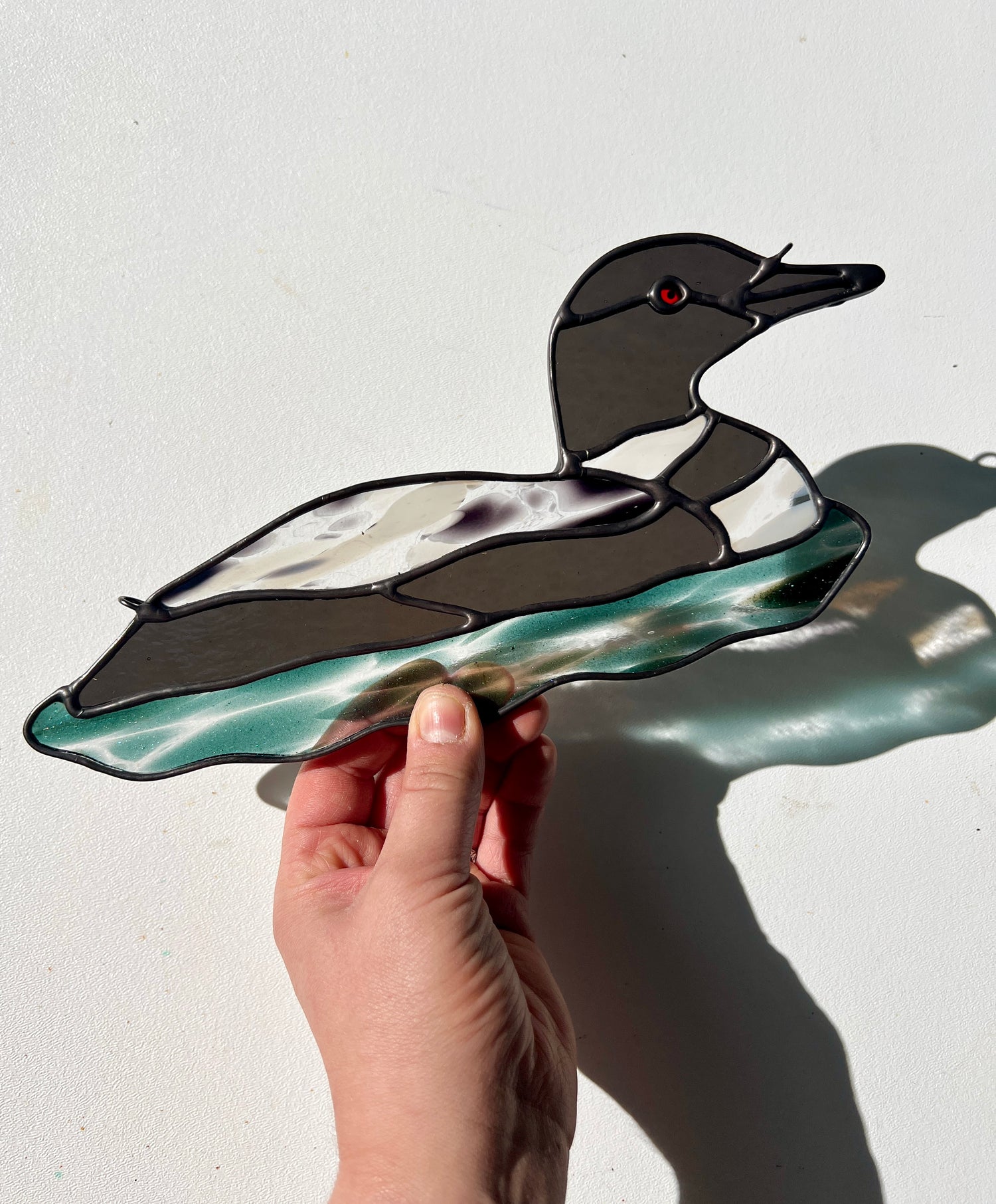 hand holding a stained glass loon over a white surface with bright sunlight casting a refraction