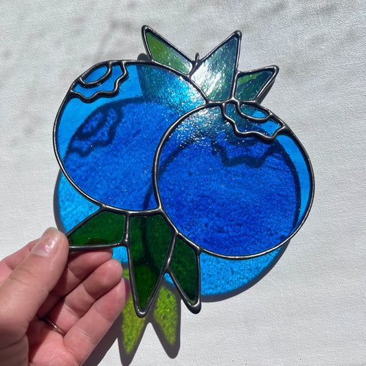 A hand holding a stained glass suncatcher depicting two blueberries and some leaves at an angle against a white background.