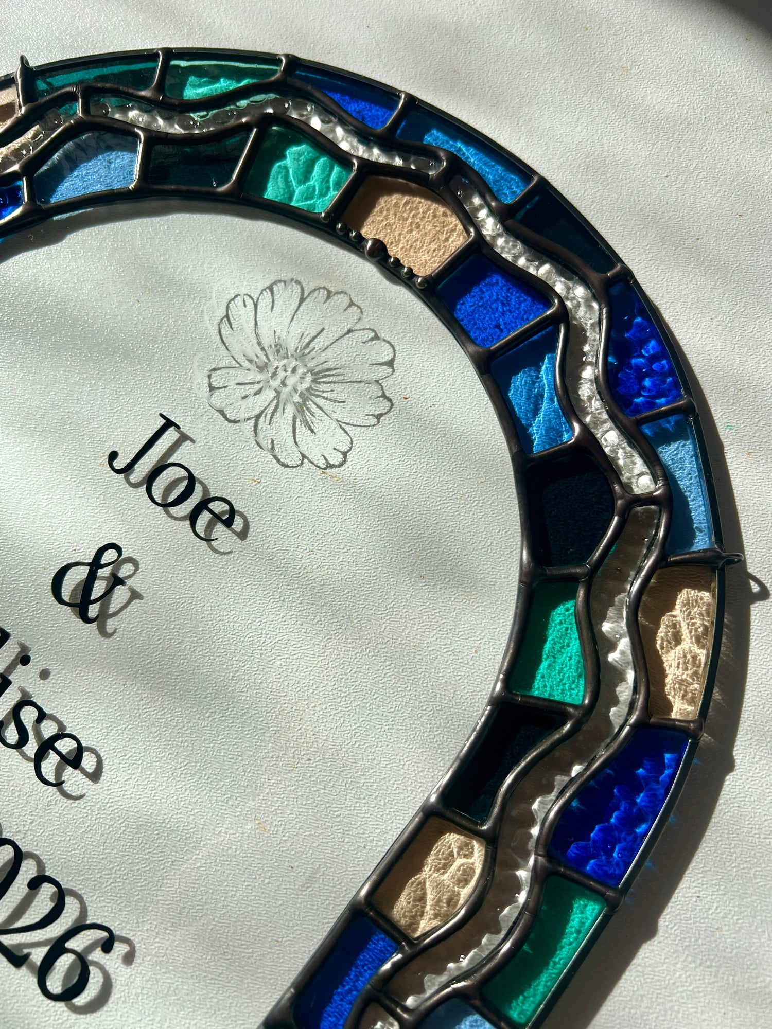 Close up of a stained glass panel with a colorful blue and green border, personalized hand painted text, and hand etched illustration of a cosmos flower.