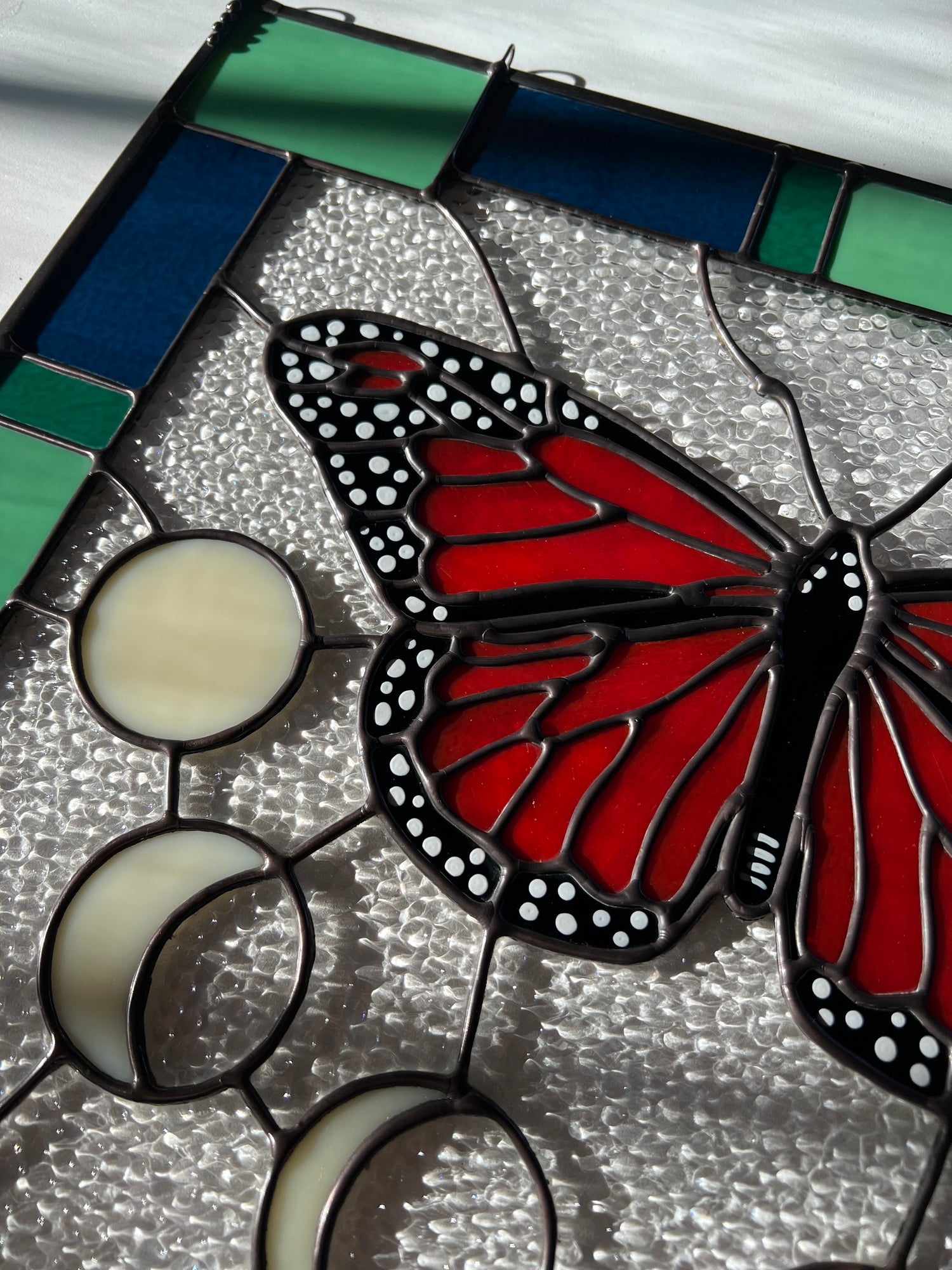 A large stained glass panel with a thick blue and green geometric border and a monarch butterfly and moon phases in the center, with textured clear glass in the background. The image provides a close up of the left half of the design, and is photographed at an angle.