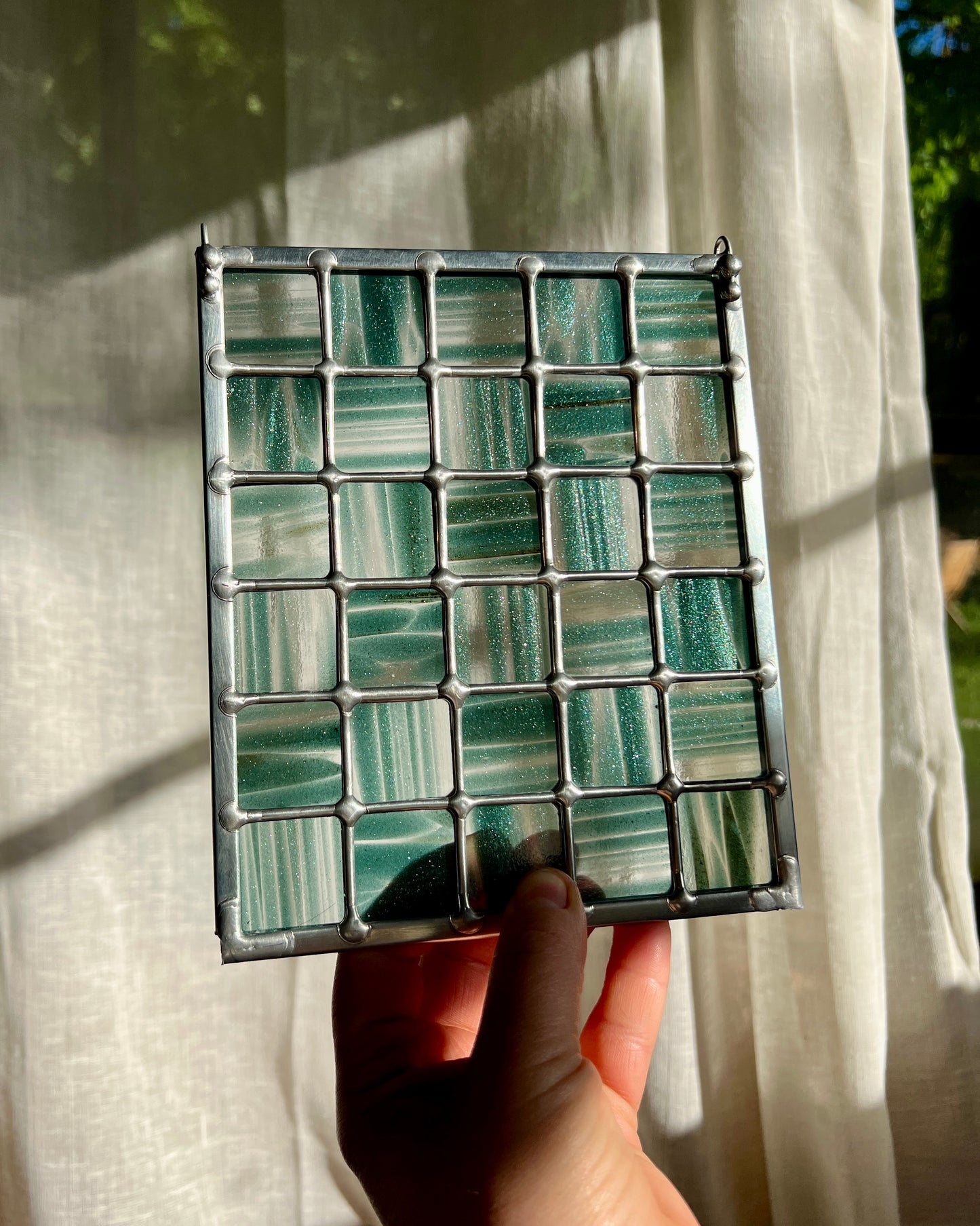 hand holding a stained glass suncatcher depicting streaky and mottled sparkly clear and teal glass arranged in a grid pattern, held in front of a window