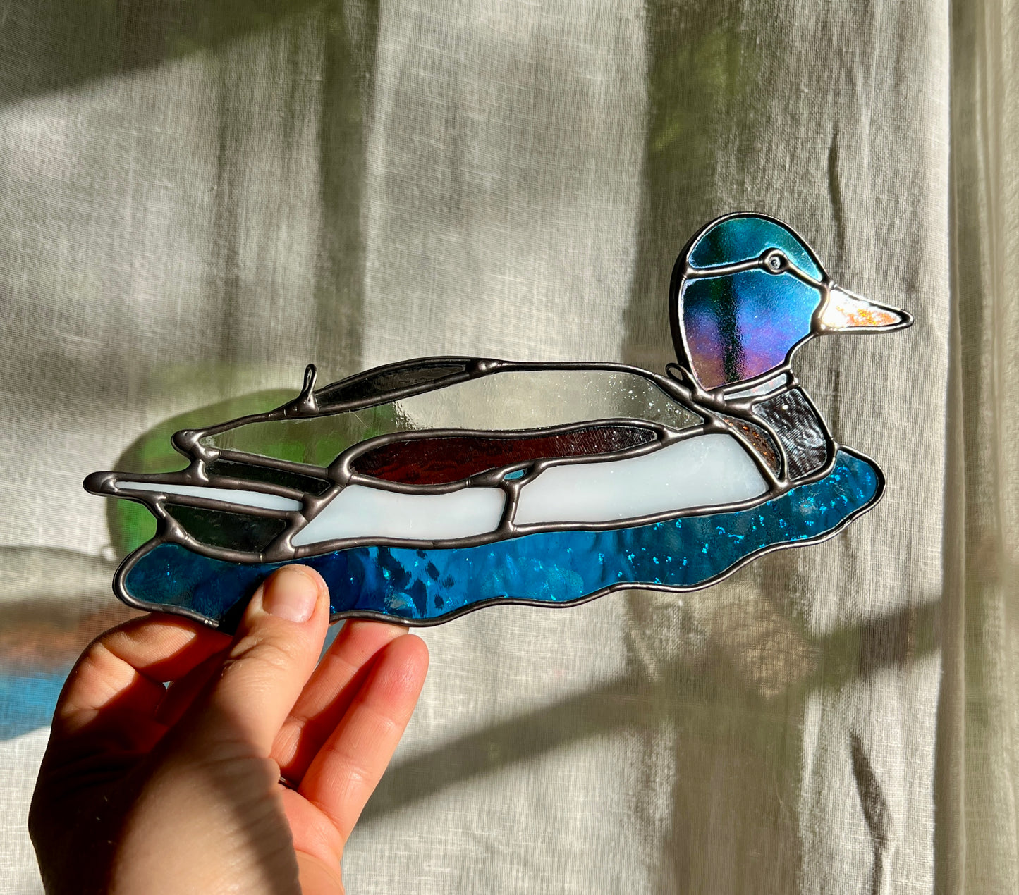 stained glass suncatcher of a male mallard held by a hand at an angle to demonstrate purple and blue iridescence of the glass used for the head