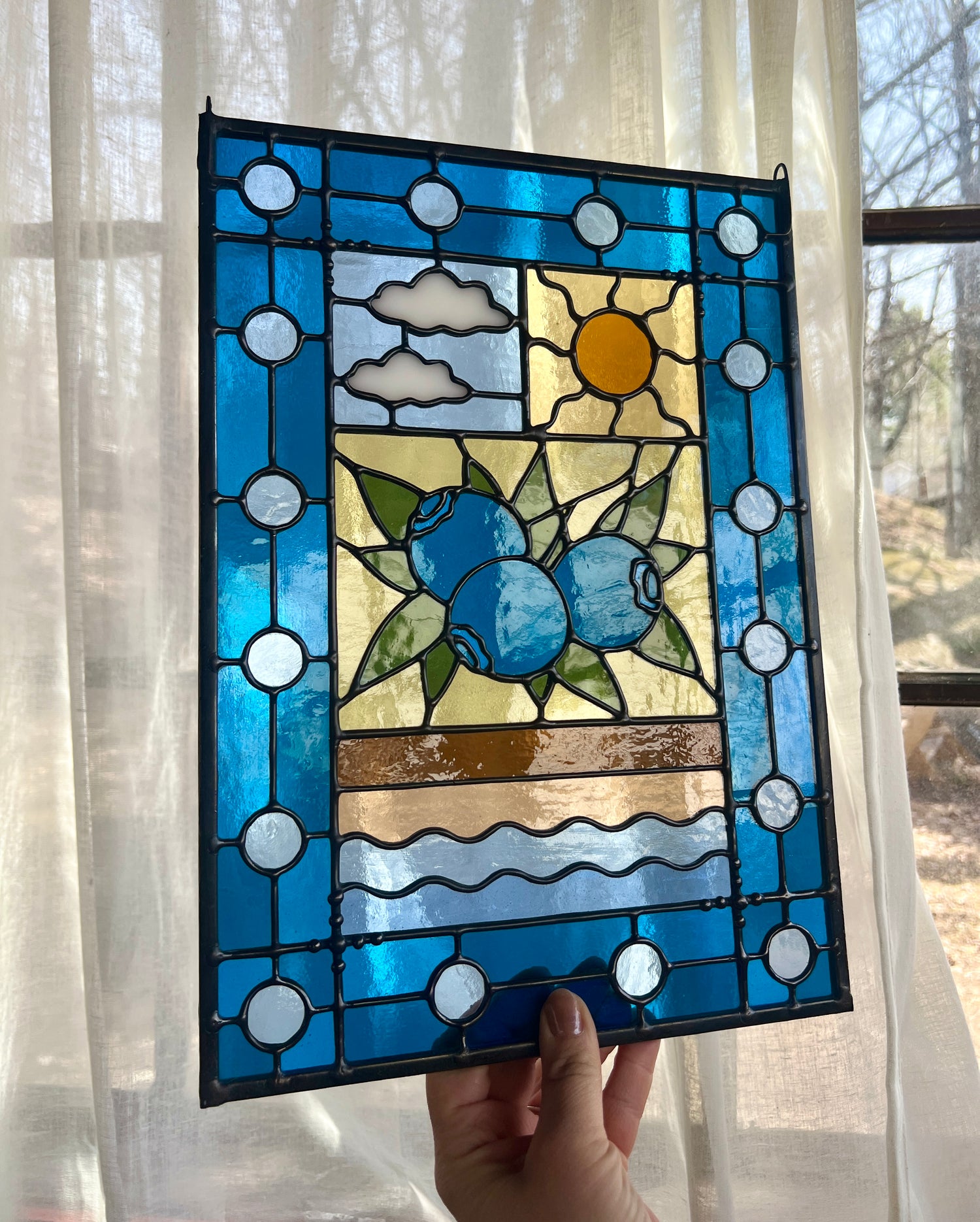 stained glass panel with a simple compartment style design featuring clouds, sun, water, and a small bunch of blueberries in the middle with a blue border, held by a hand at an angle in front of a window