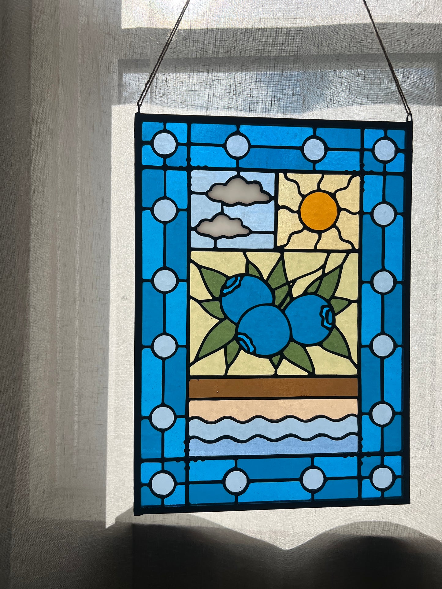 stained glass panel with a simple compartment style design featuring clouds, sun, water, and a small bunch of blueberries in the middle with a blue border