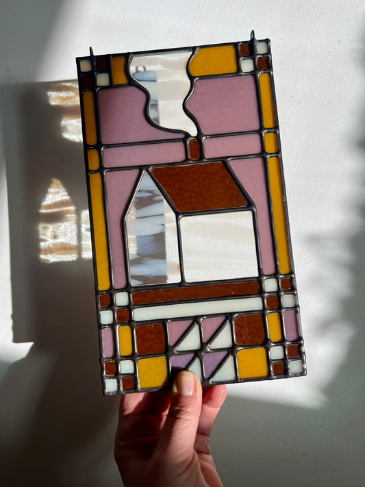 Stained glass textile inspired panel depicting a home in the center in shades of purple, white, brown, and bronze, held by a hand above a white surface with sunlight casting subtle refractions
