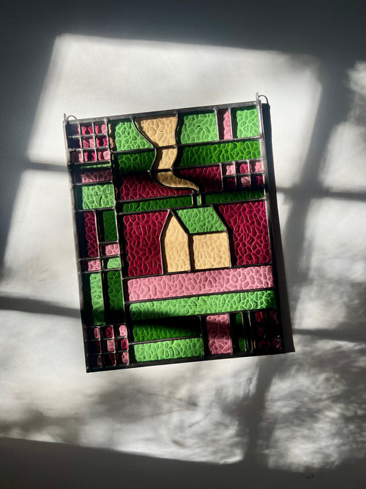 Stained glass panel with geometric textile inspired design with a cozy home depicted in the center, laying on a white black ground with bright sunlight across it, showing off the textured glass