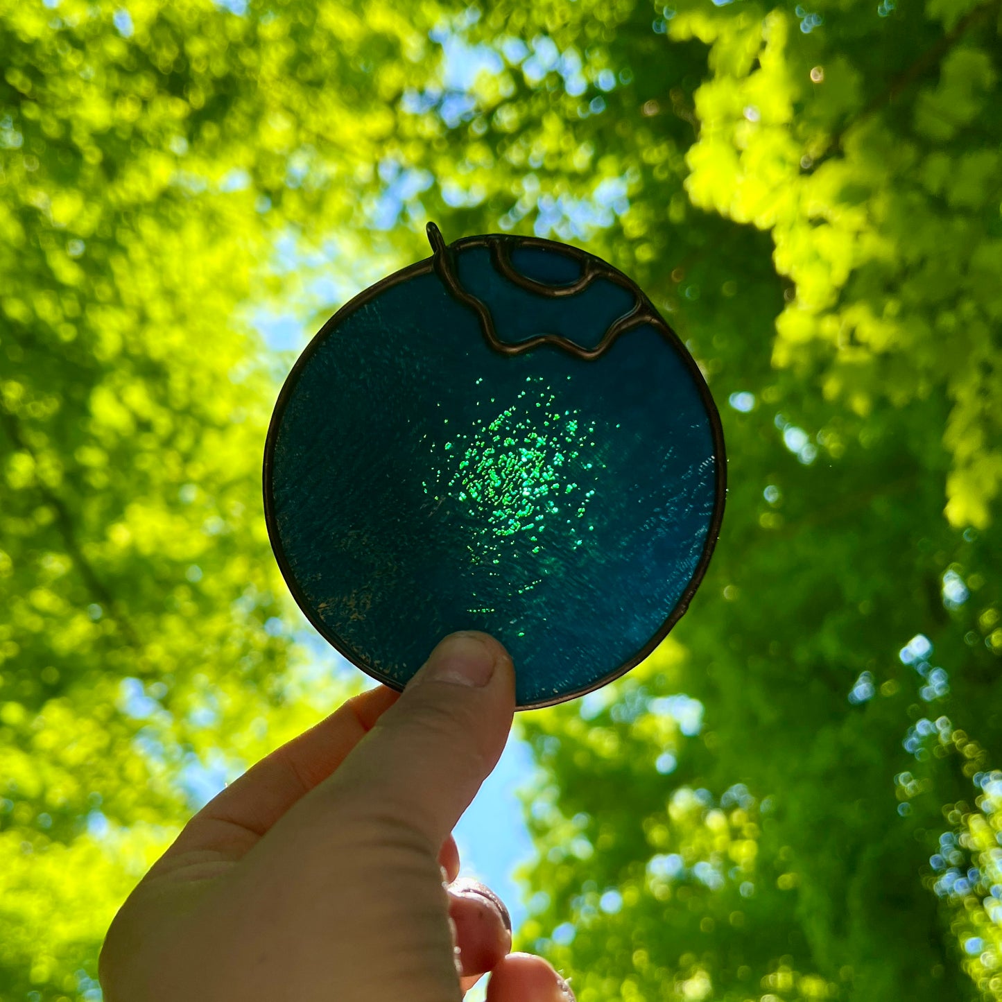 photo of a stained glass blueberry held by a hand up to sunlight with bright green leaves and trees in the background