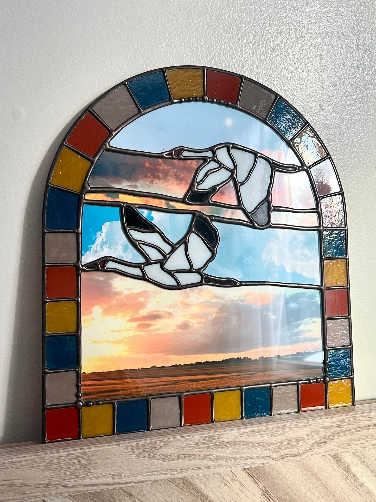 Stained glass panel with a colorful border featuring two birds and a sunset scene on a white wall.