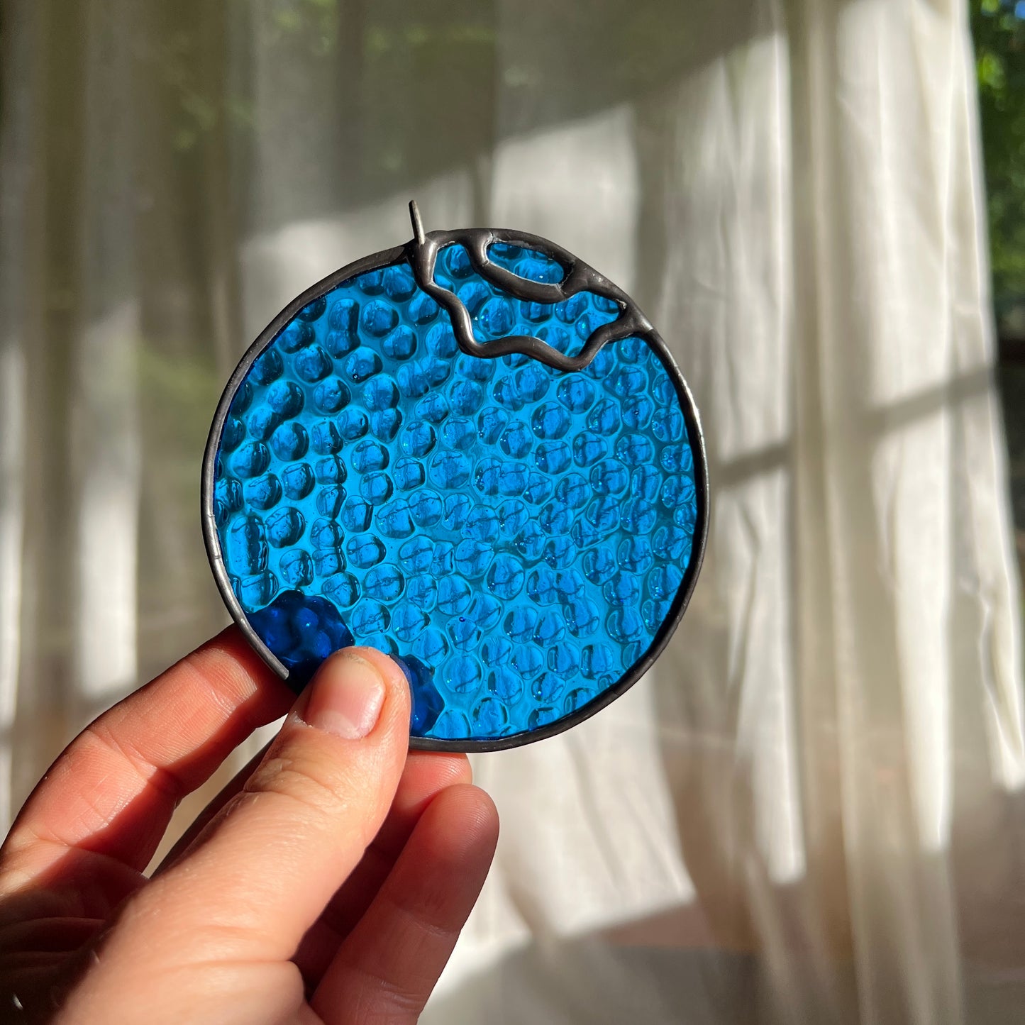 hand holding a textured stained glass blueberry in front of a window