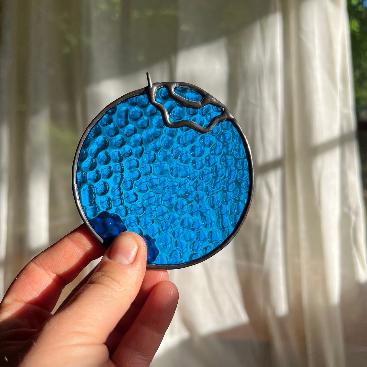 hand holding a textured stained glass blueberry in front of a window