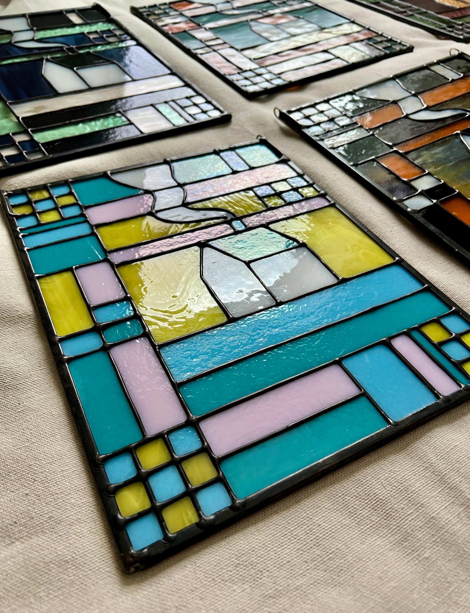 Stained glass textile inspired panel depicting a home in the center in shades of teal, pink, white, blue, and yellow, photographed at an angle to show smooth soldering details.