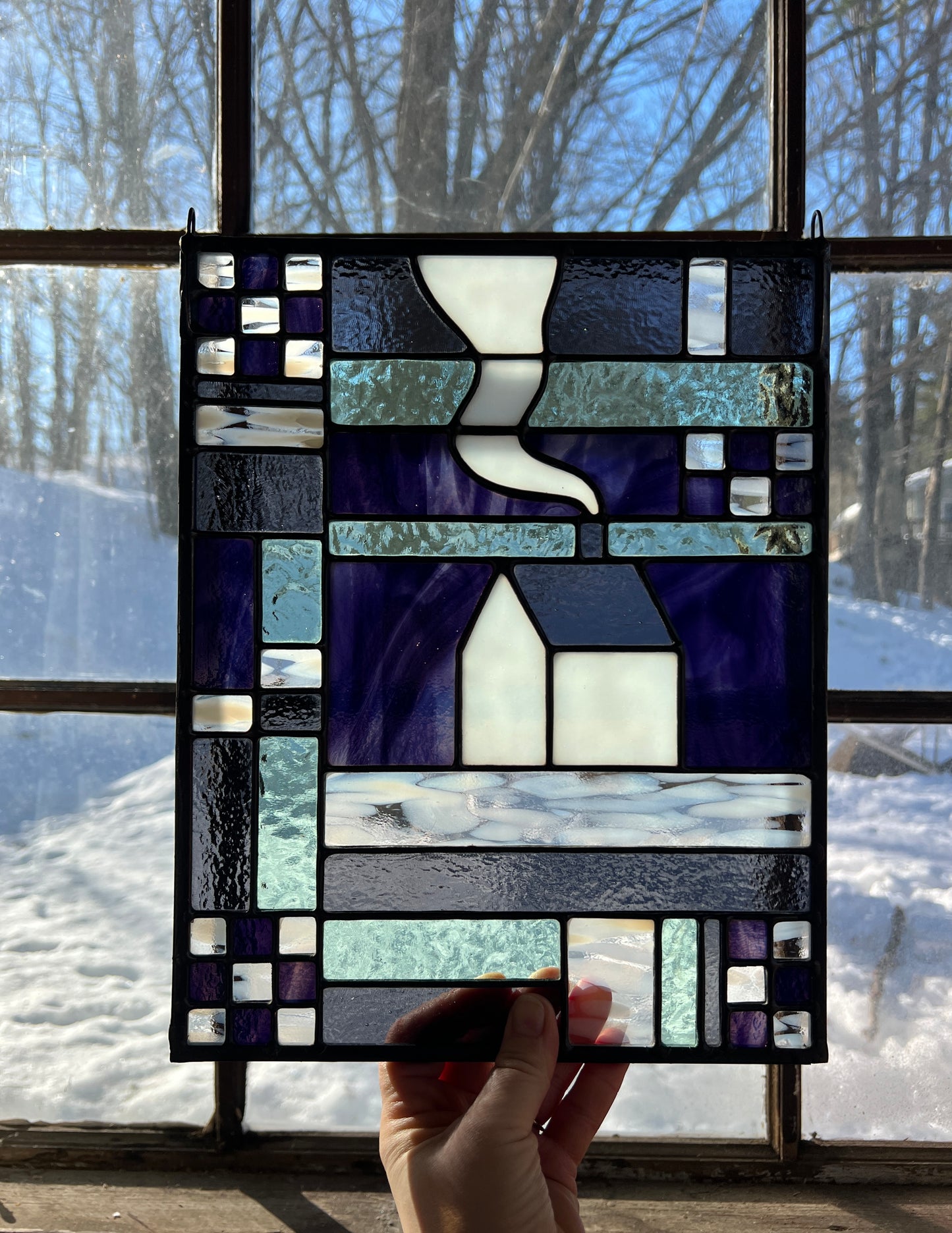 Stained glass panel with geometric textile inspired design with a cozy home depicted in the center, held by a hand in front of a window with a snowy backdrop.