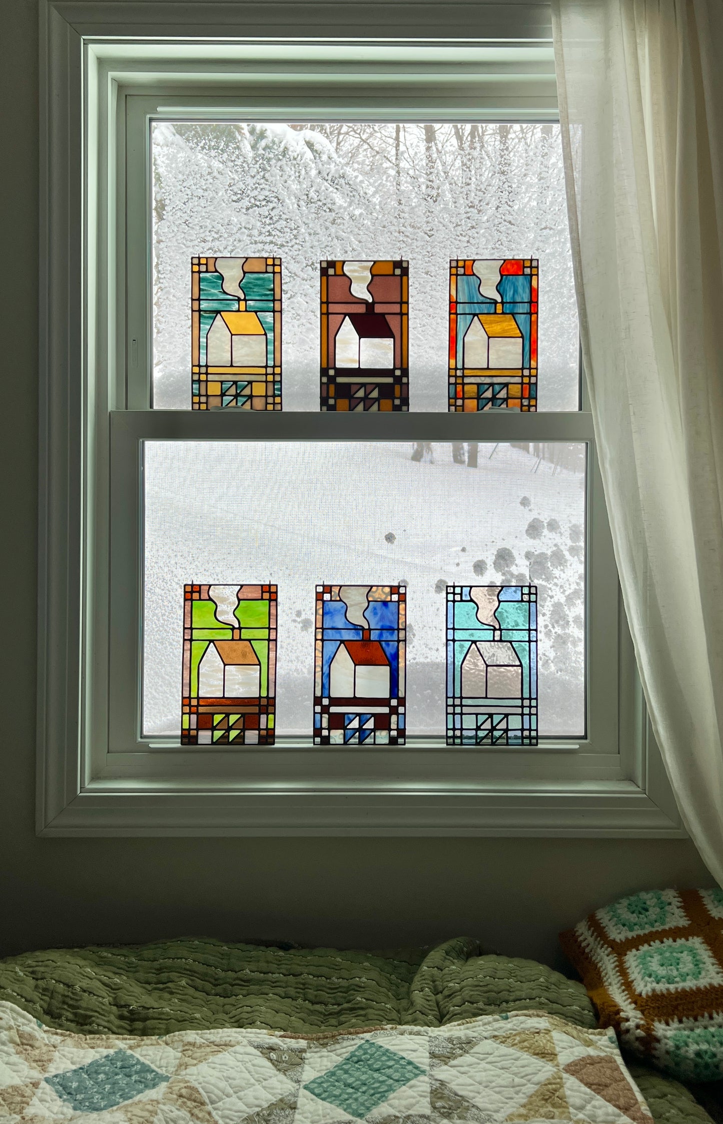 A set of six textile inspired stained glass panels in various color combinations resting in a window with a snow scene outside and cozy blankets in the foreground.