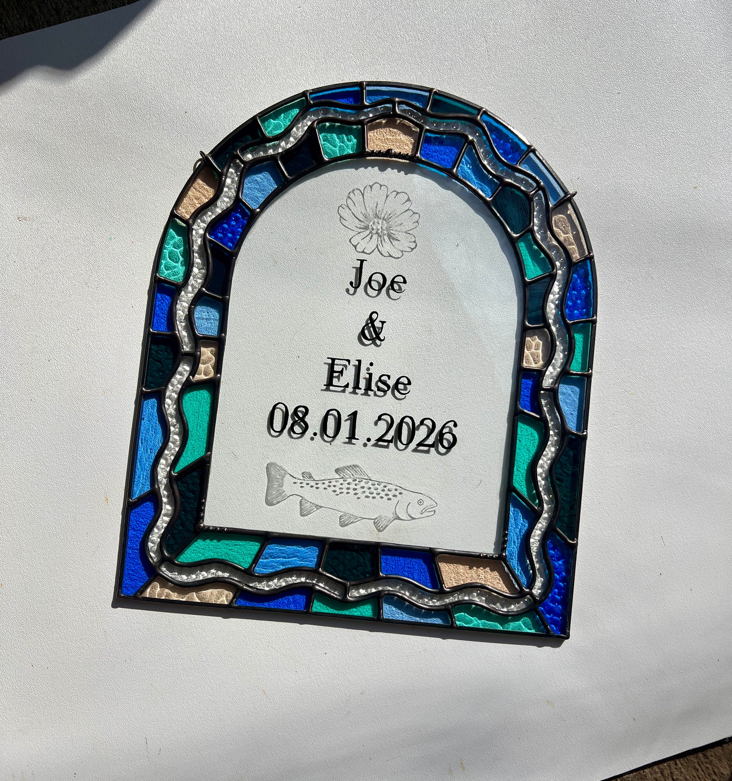 Stained glass panel in the shape of an arch with a multicolor blue, teal, turquoise, and pink border. Within the border is squiggle detail in clear textured glass. The center of the arch is clear uncolored glass, with personalized text and hand etched details of a cosmos flower and trout. The piece is held resting against a white background with bright sunlight cast across it.