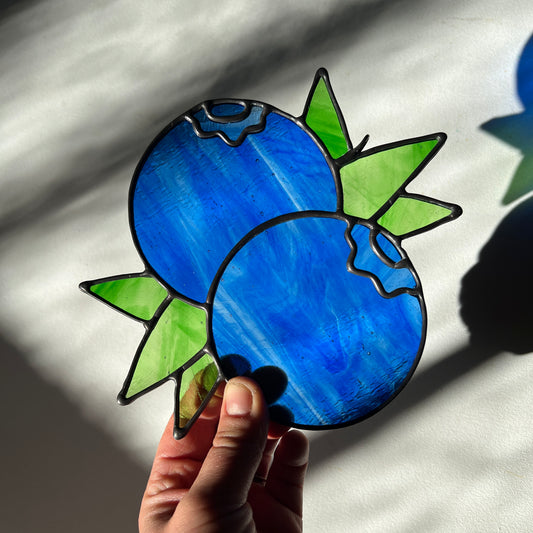 hand holding a wispy blue stained glass suncatcher depicting a small bunch of blueberries with leaves