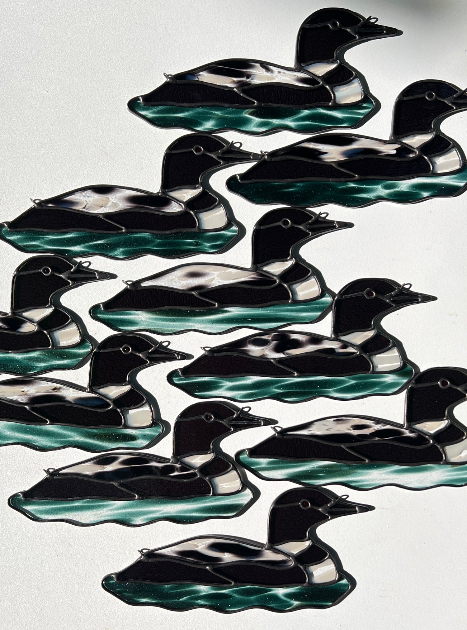 ten stained glass loons arranged in a group on a white surface with bright sunlight cast across