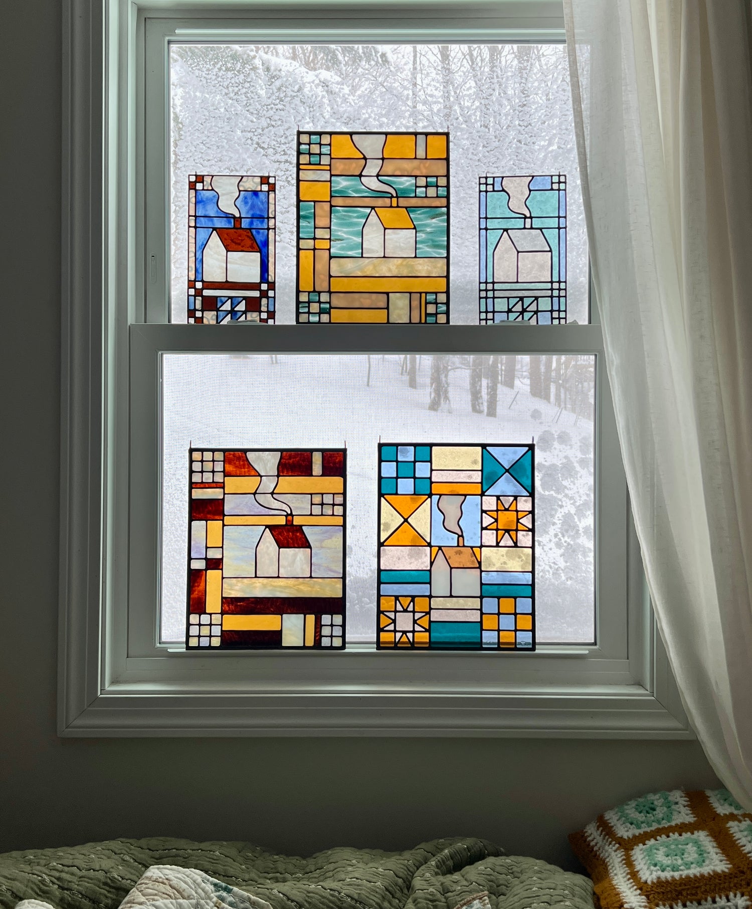 Five textile inspired stained glass panels in various color combinations resting in a window with a snowy landscape outside and cozy blankets in the foreground