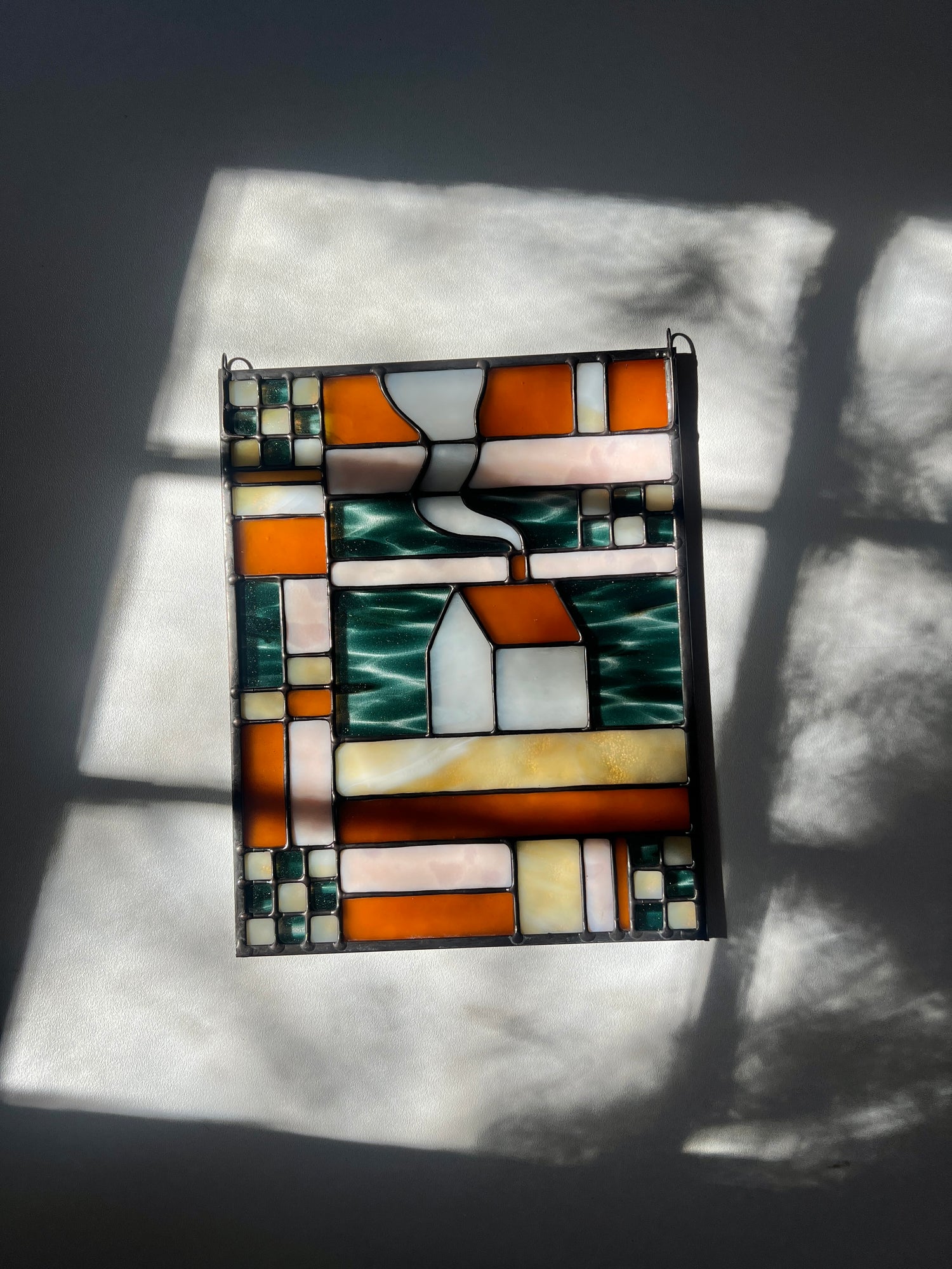 Stained glass textile inspired panel depicting a home in the center in shades of teal, amber, white, and beige, resting on a white surface with sunlight and shadows cast across