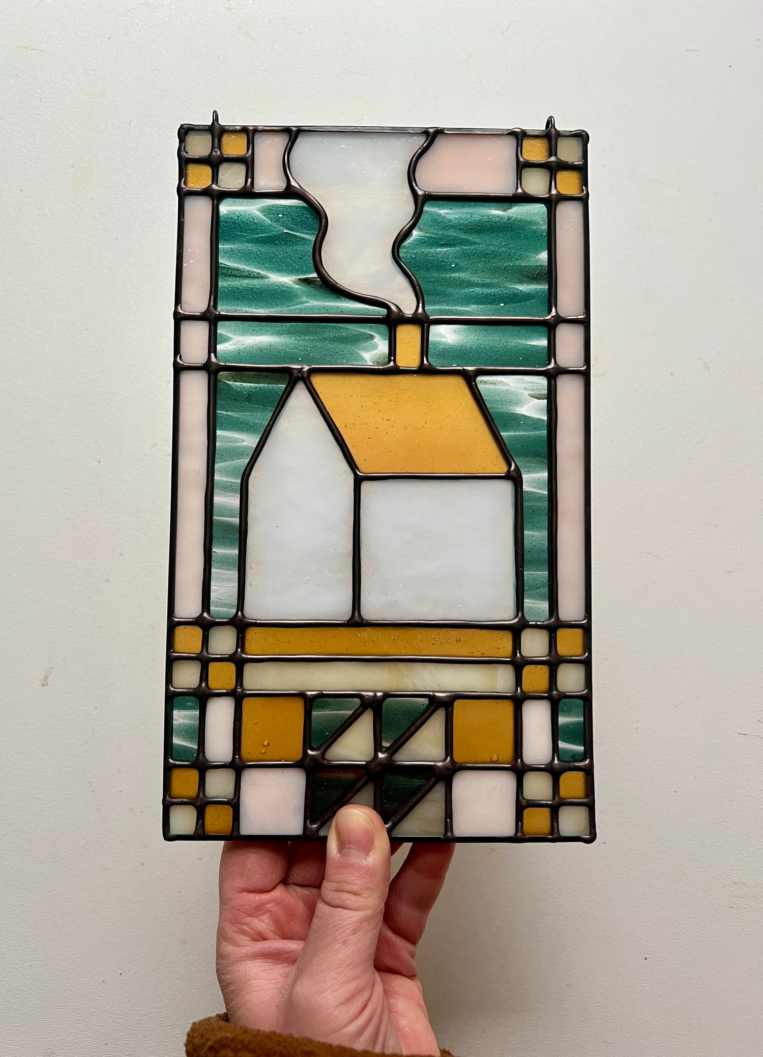 Stained glass textile inspired panel depicting a home in the center in shades of teal, amber, white, and beige, held by a hand above a white surface