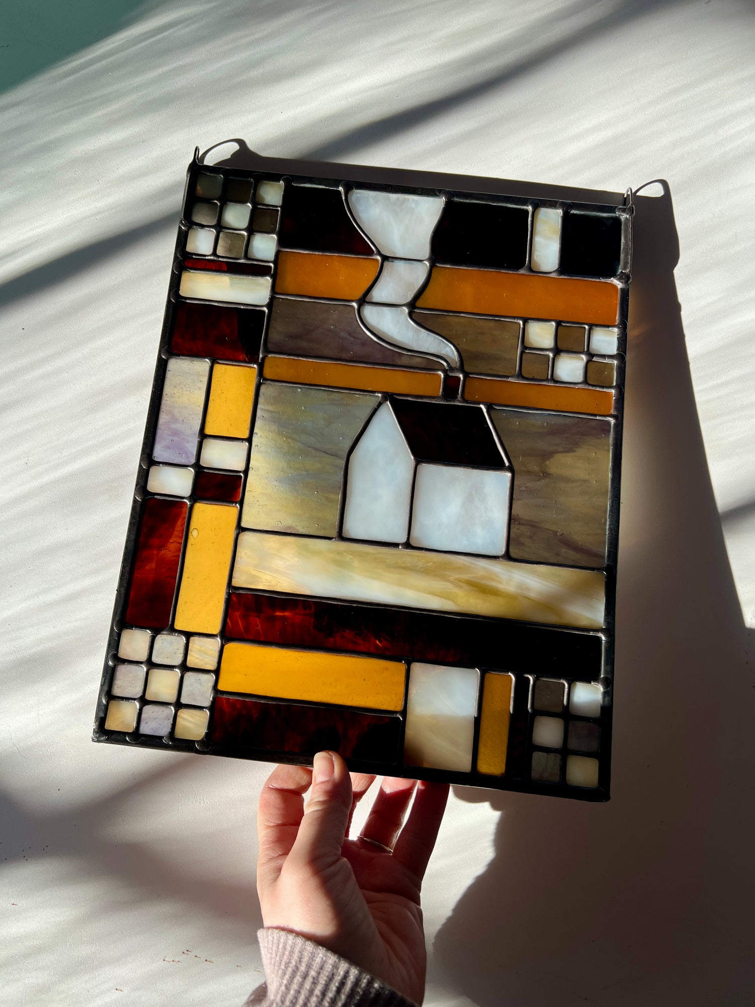 Stained glass textile inspired panel depicting a home in the center in shades of purple, white, brown, and amber, held by a hand against a white surface with sunlight and shadows cast across