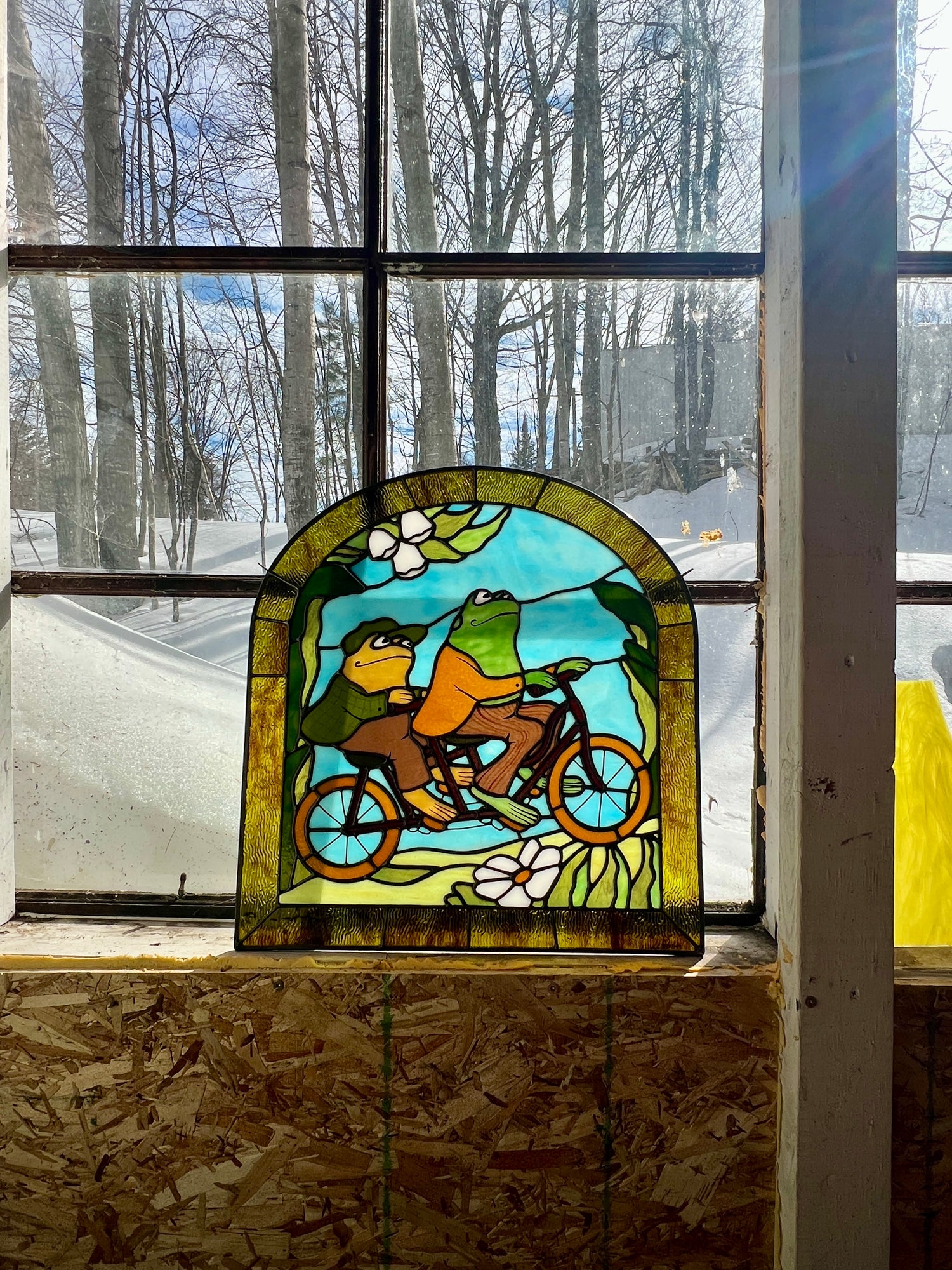stained glass panel depicting frog and toad riding a bicycle leaning against a large window with a snowy landscape outside