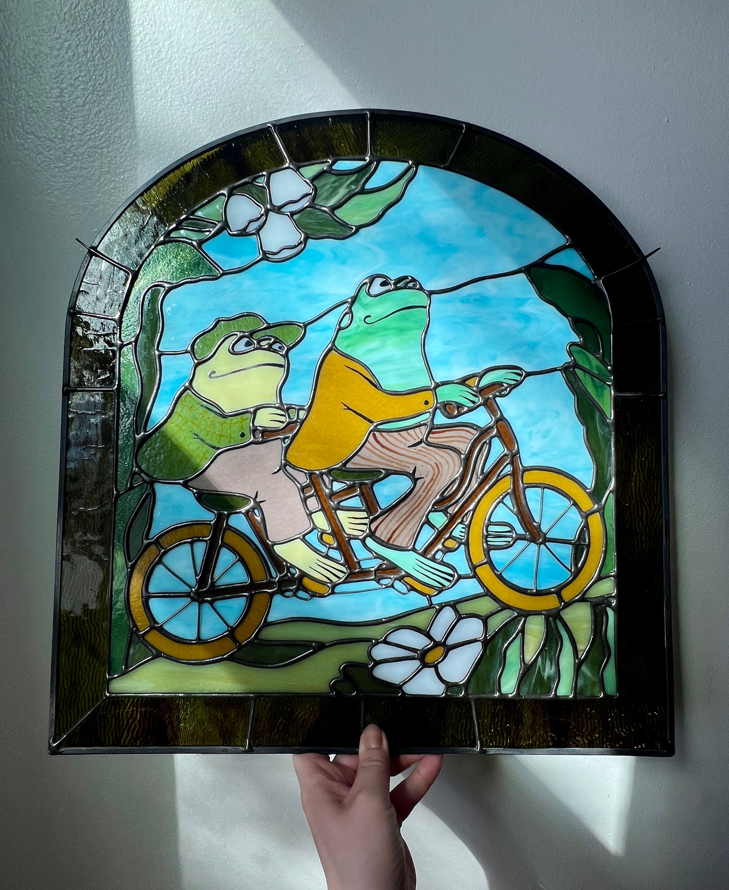 a hand holding a stained glass panel depicting frog and toad riding a bicycle resting against a white surface