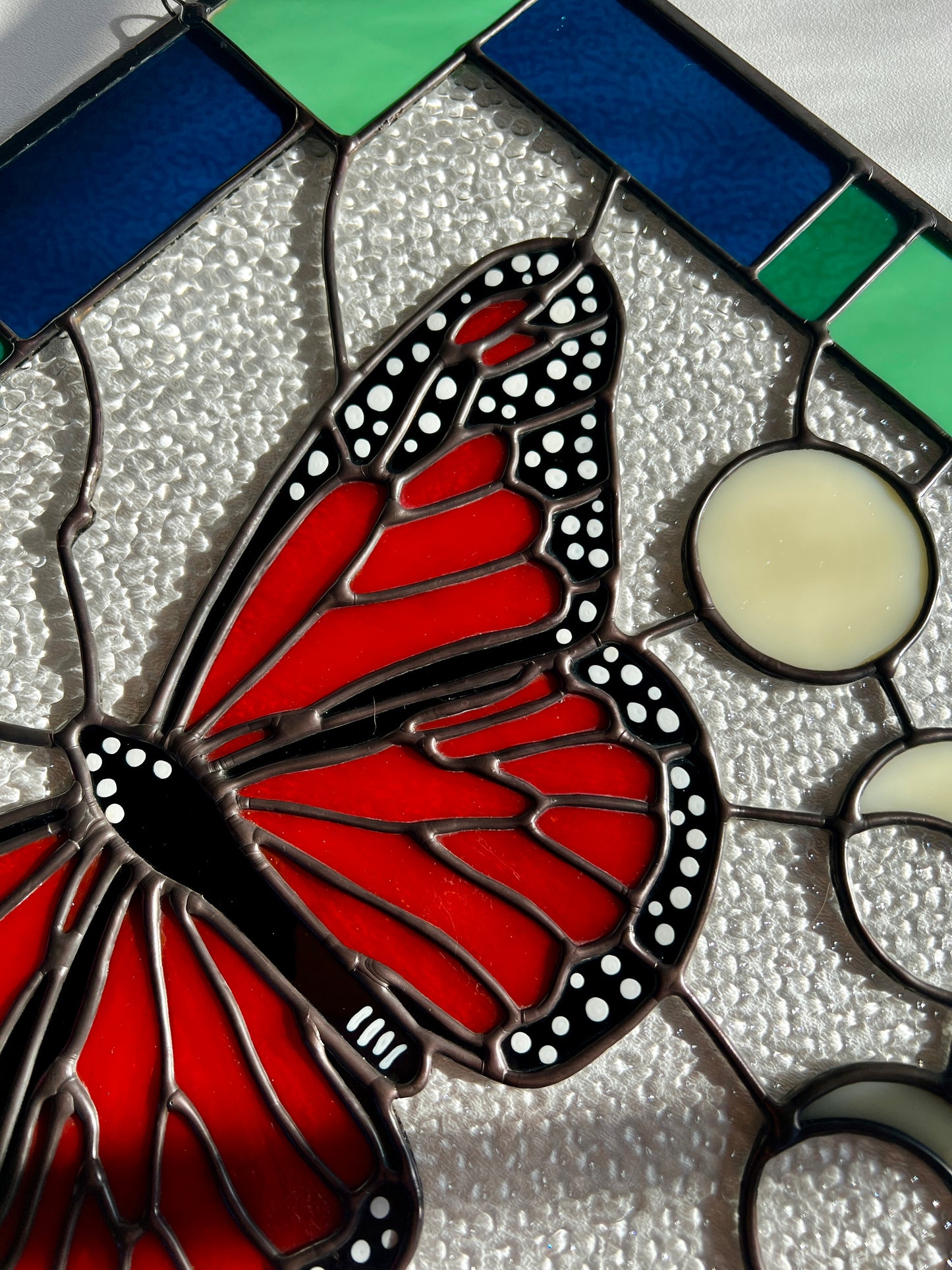 Close up up hand painted details on a large stained glass panel with a thick blue and green geometric border and a monarch butterfly and moon phases in the center, with textured clear glass in the background.