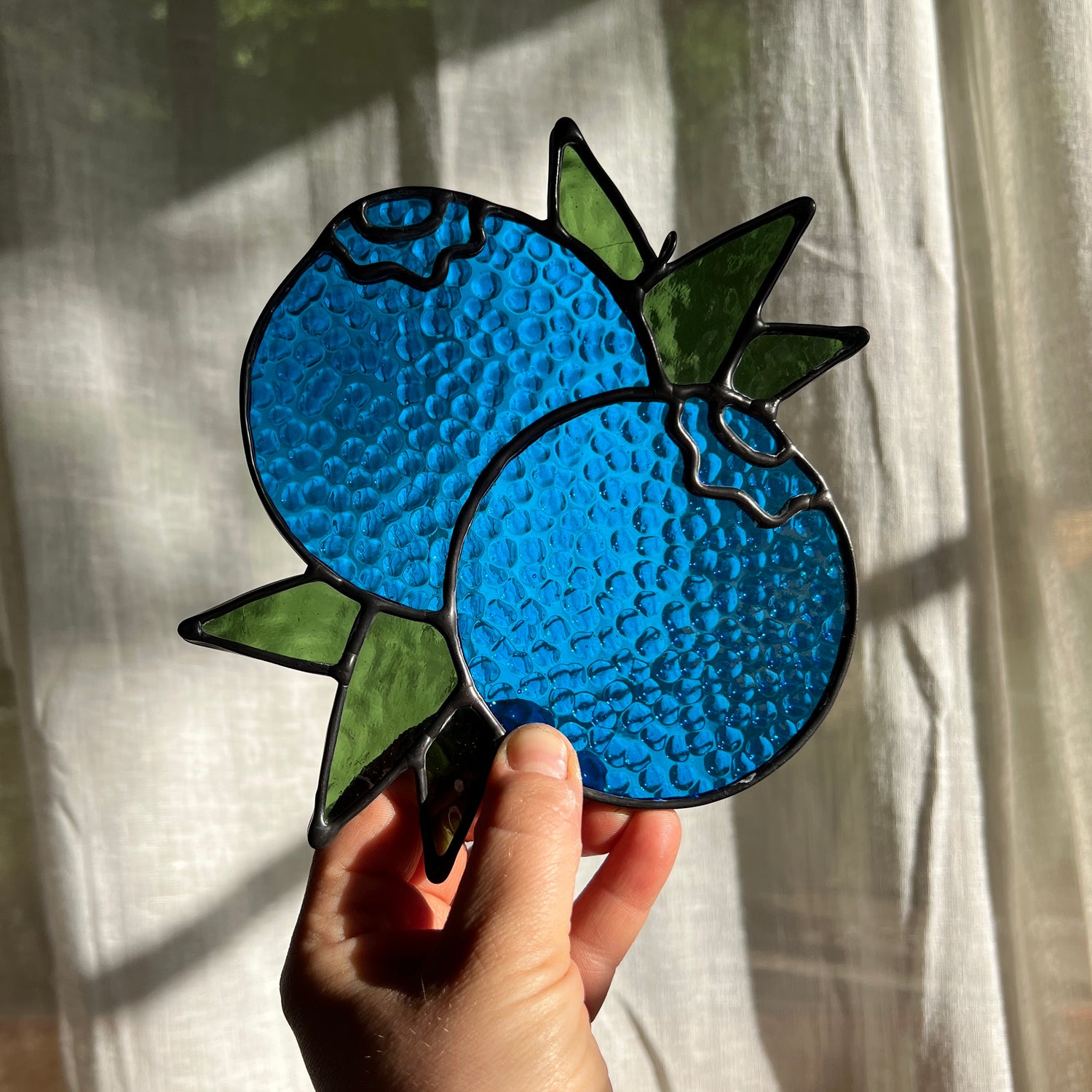 hand holding a textured stained glass suncatcher depicting a small bunch of blueberries with leaves in front of a window