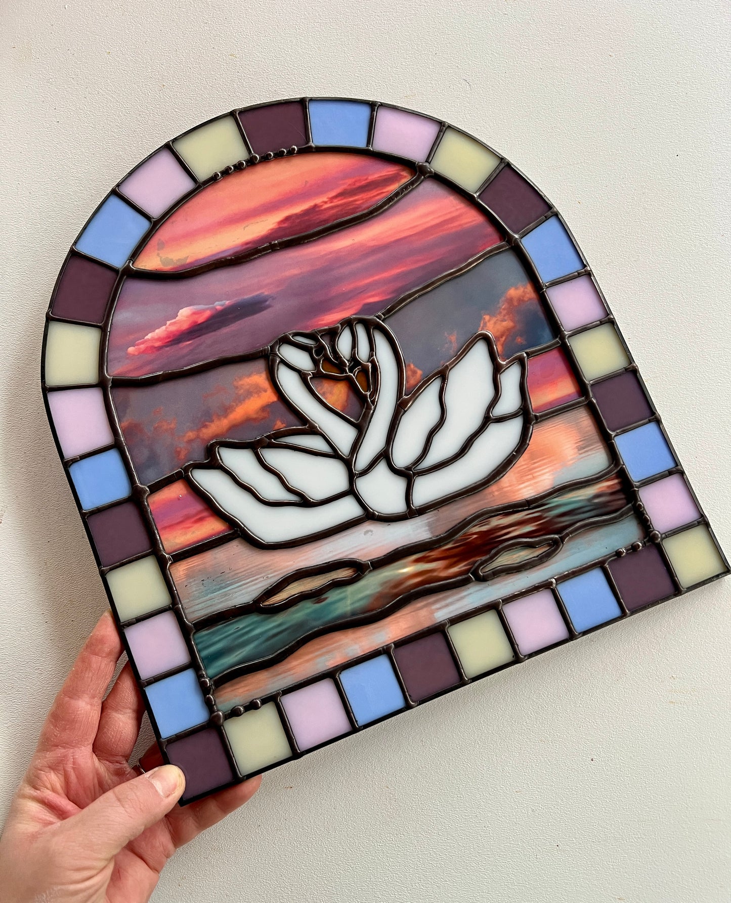 Stained glass panel with a colorful pastel arch border and two white swans in the center. Collaged photographs are used to create a sunset background.