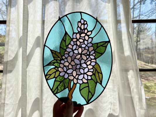 Stained glass panel with floral lilac design held in front of a window with sheer curtains.