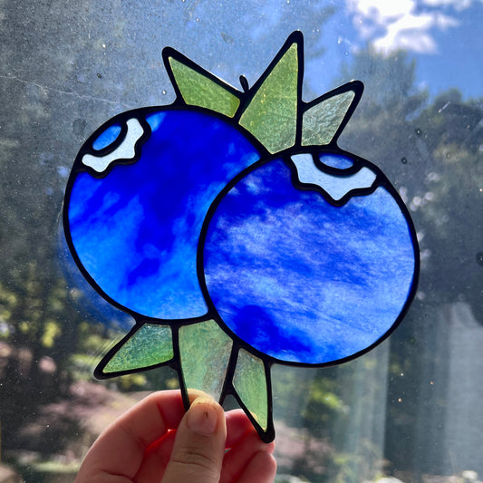 A hand holding a stained glass suncatcher depicting two blueberries and some leaves against a window with backlighting.
