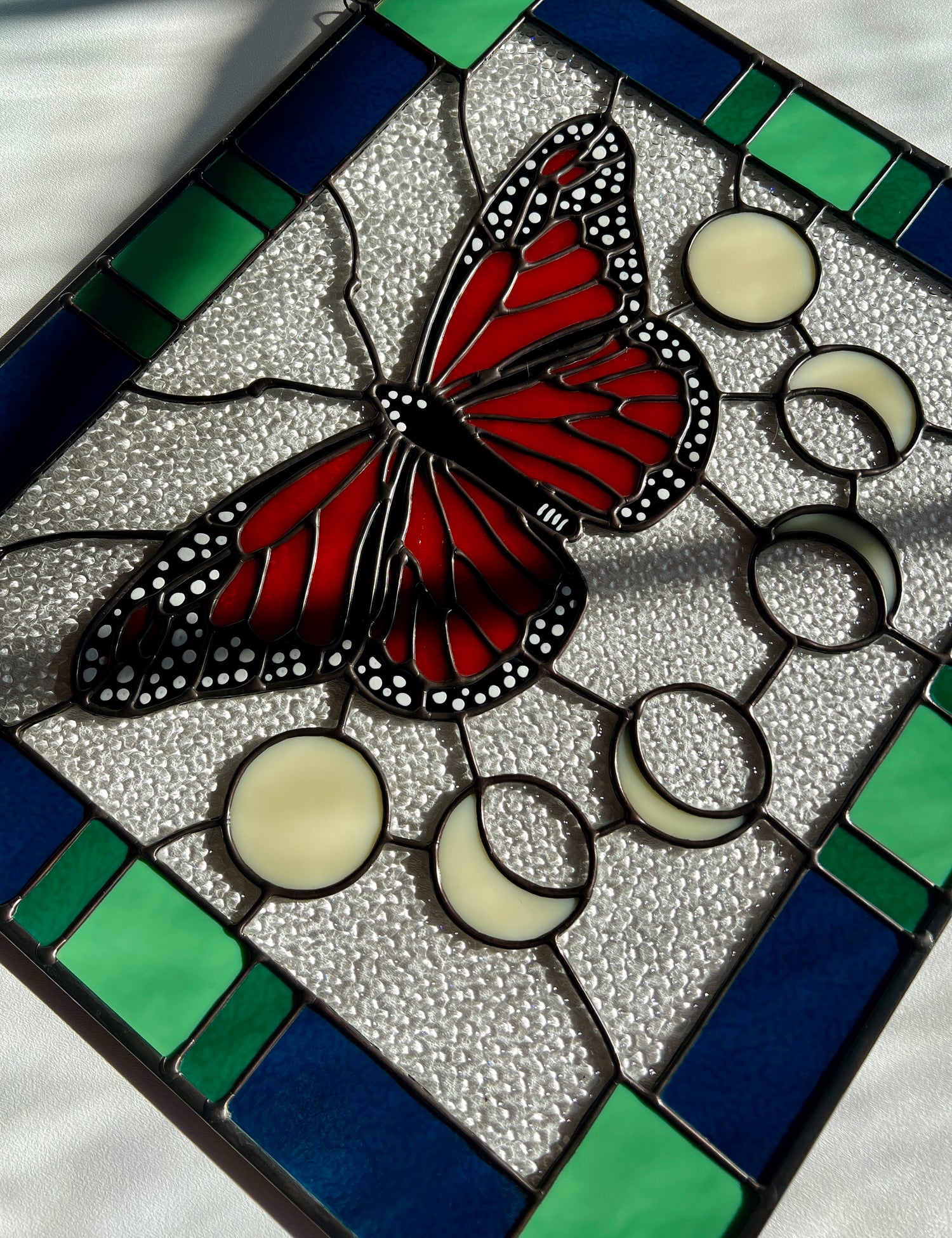 A large stained glass panel with a thick blue and green geometric border and a monarch butterfly and moon phases in the center, with textured clear glass in the background. The piece is photographed at an angle with contrasting sunlight and shadows falling across it.