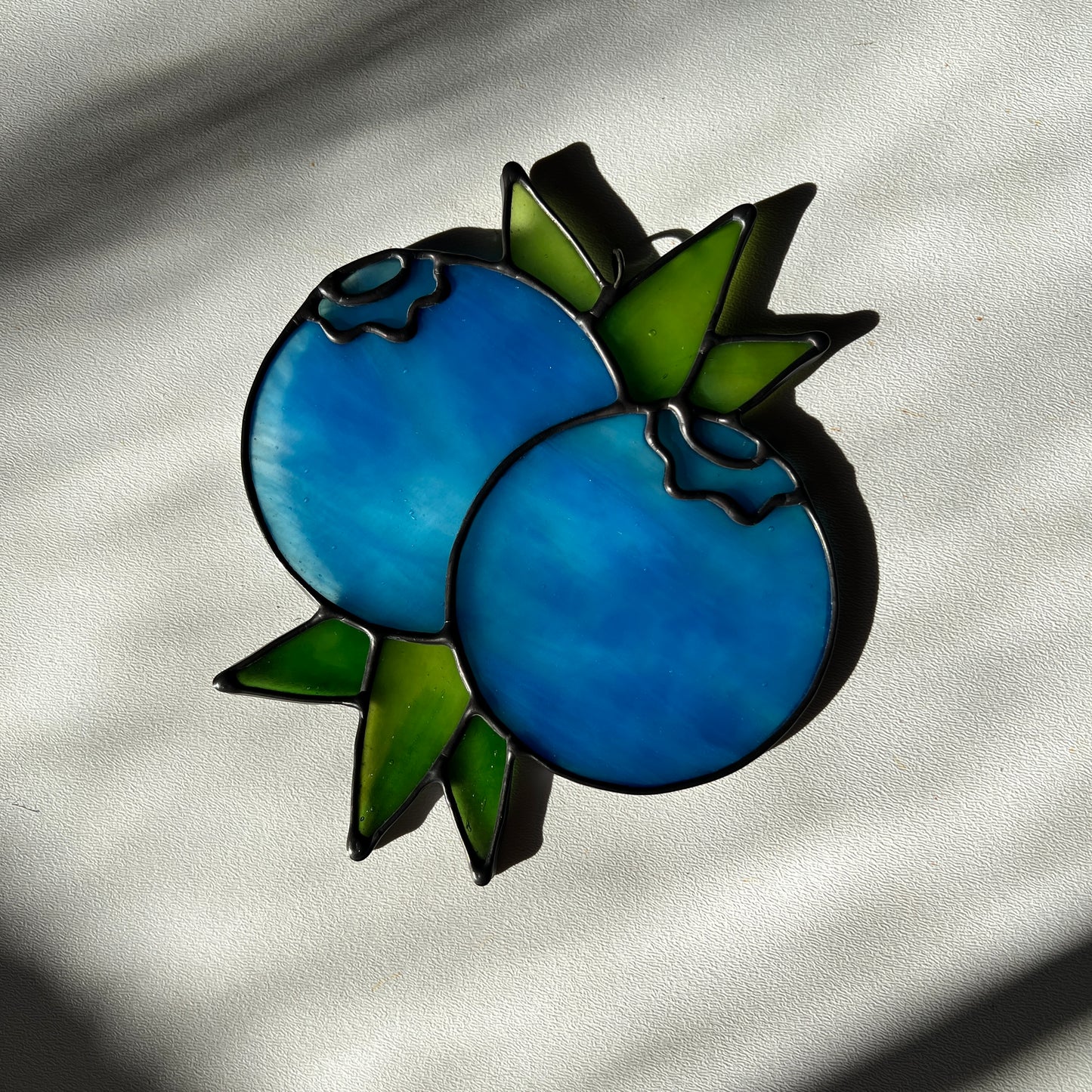 a  wispy light blue stained glass suncatcher depicting a small bunch of blueberries with leaves resting on a white surface