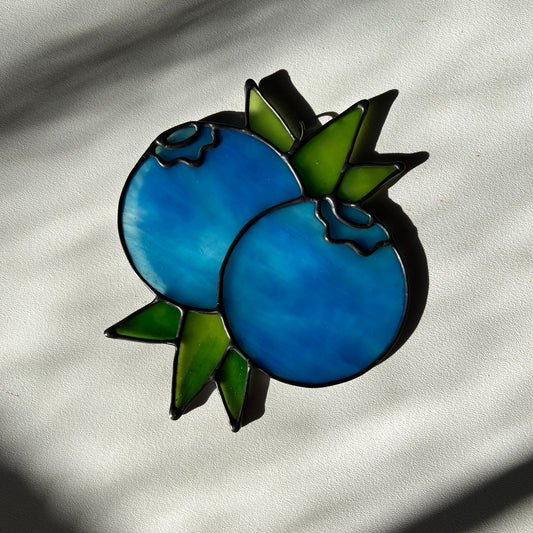 a  wispy light blue stained glass suncatcher depicting a small bunch of blueberries with leaves resting on a white surface