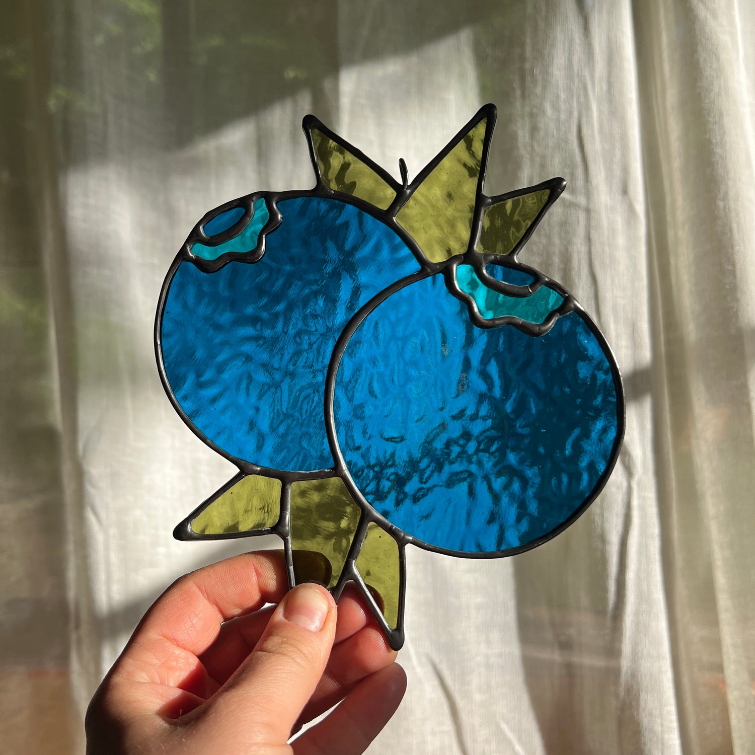 hand holding a textured blue stained glass suncatcher depicting a small bunch of blueberries with leaves in front of a window