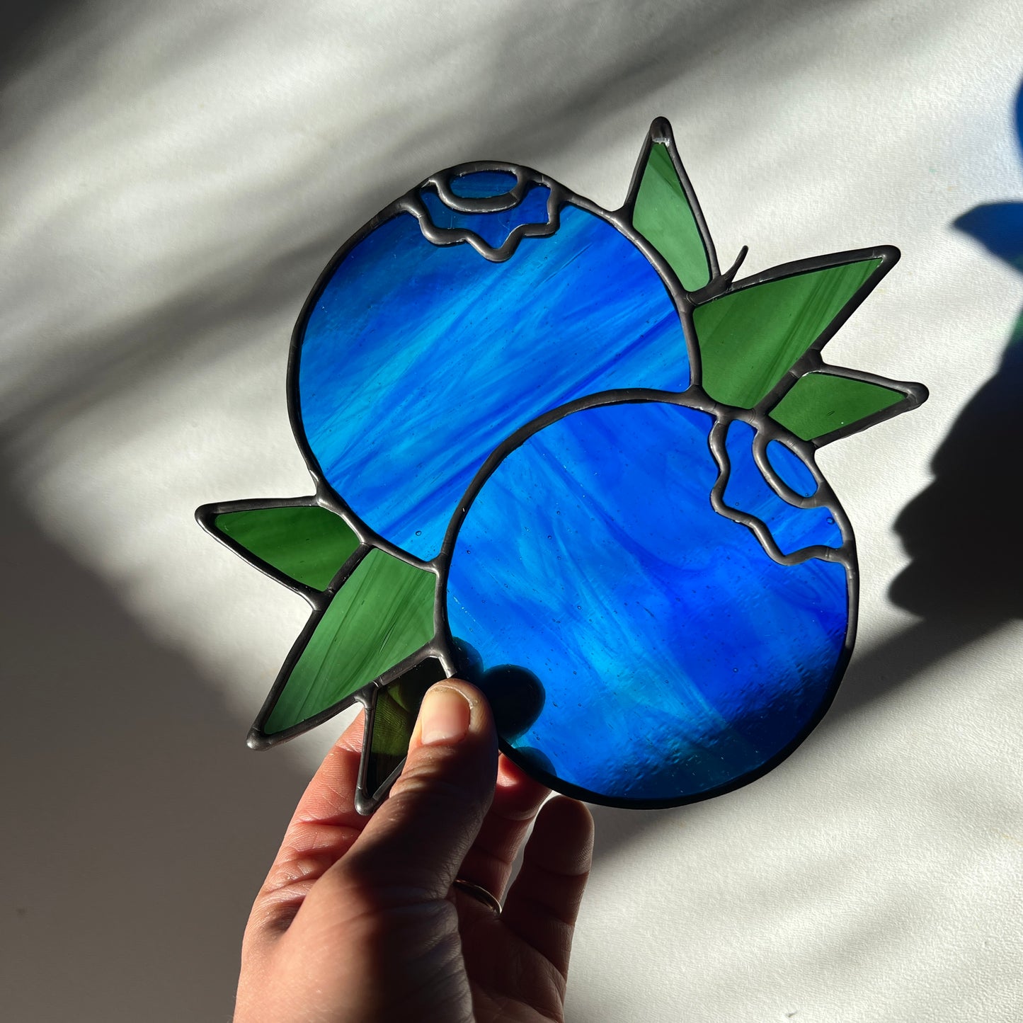 hand holding a wispy blue stained glass suncatcher depicting a small bunch of blueberries with leaves